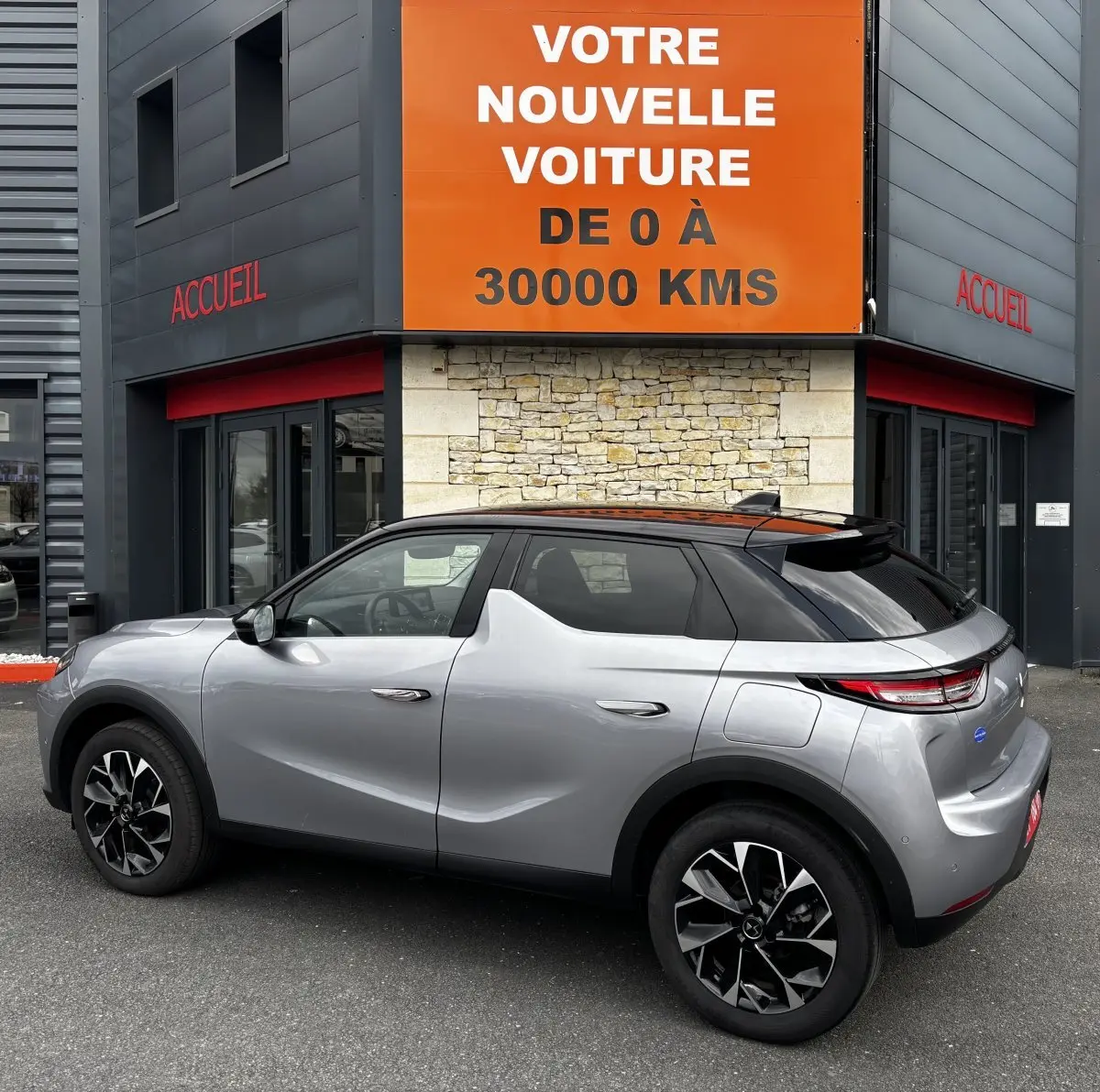 Vue 3/4 arrière droite d'une DS3 gris clair avec toit noir, jantes noires et détails modernes, devant un bâtiment commercial.