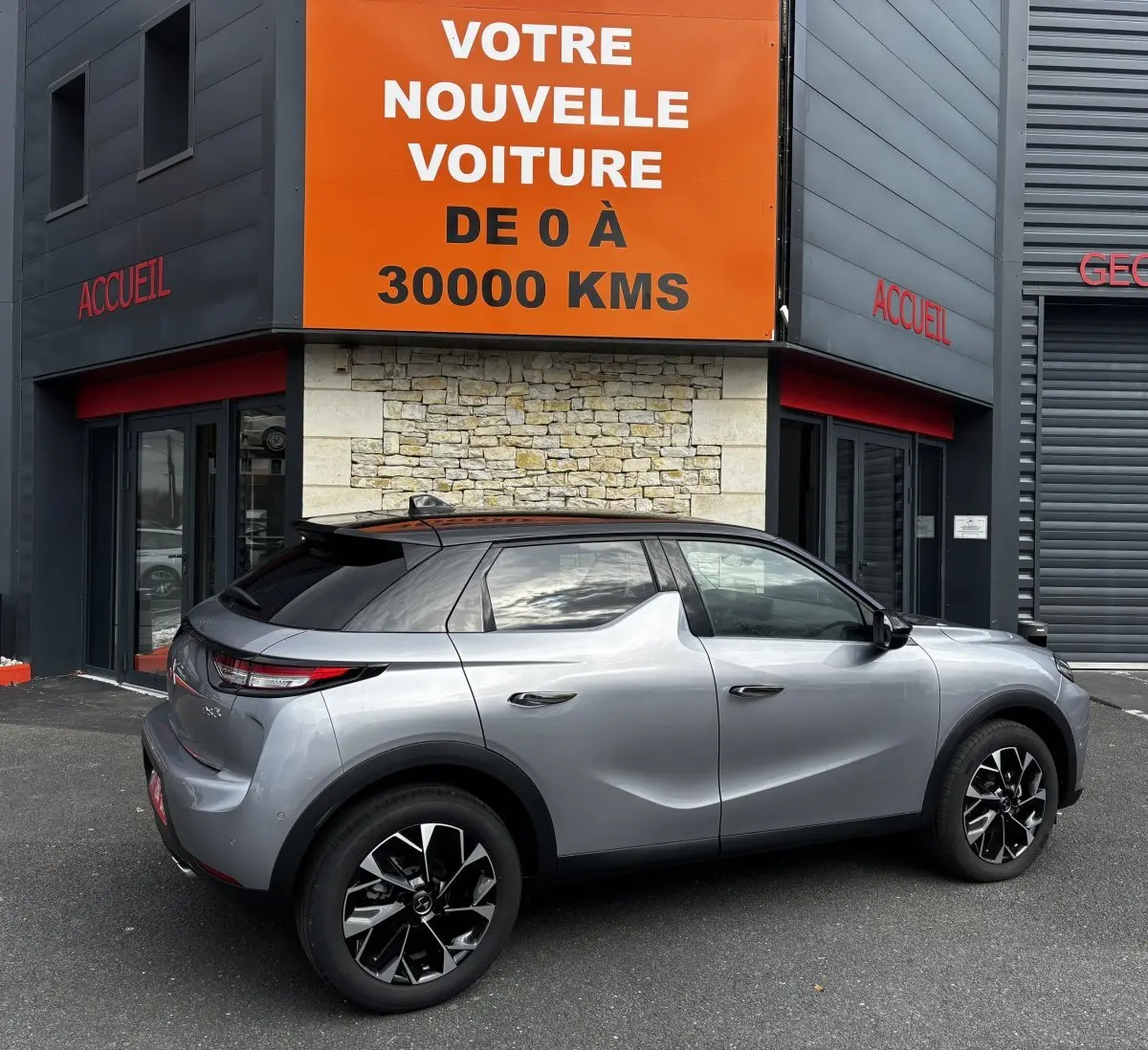 Vue latérale droite d'une DS3 2024 gris clair avec toit noir, jantes alliage noires et vitres teintées, garée devant un bâtiment.