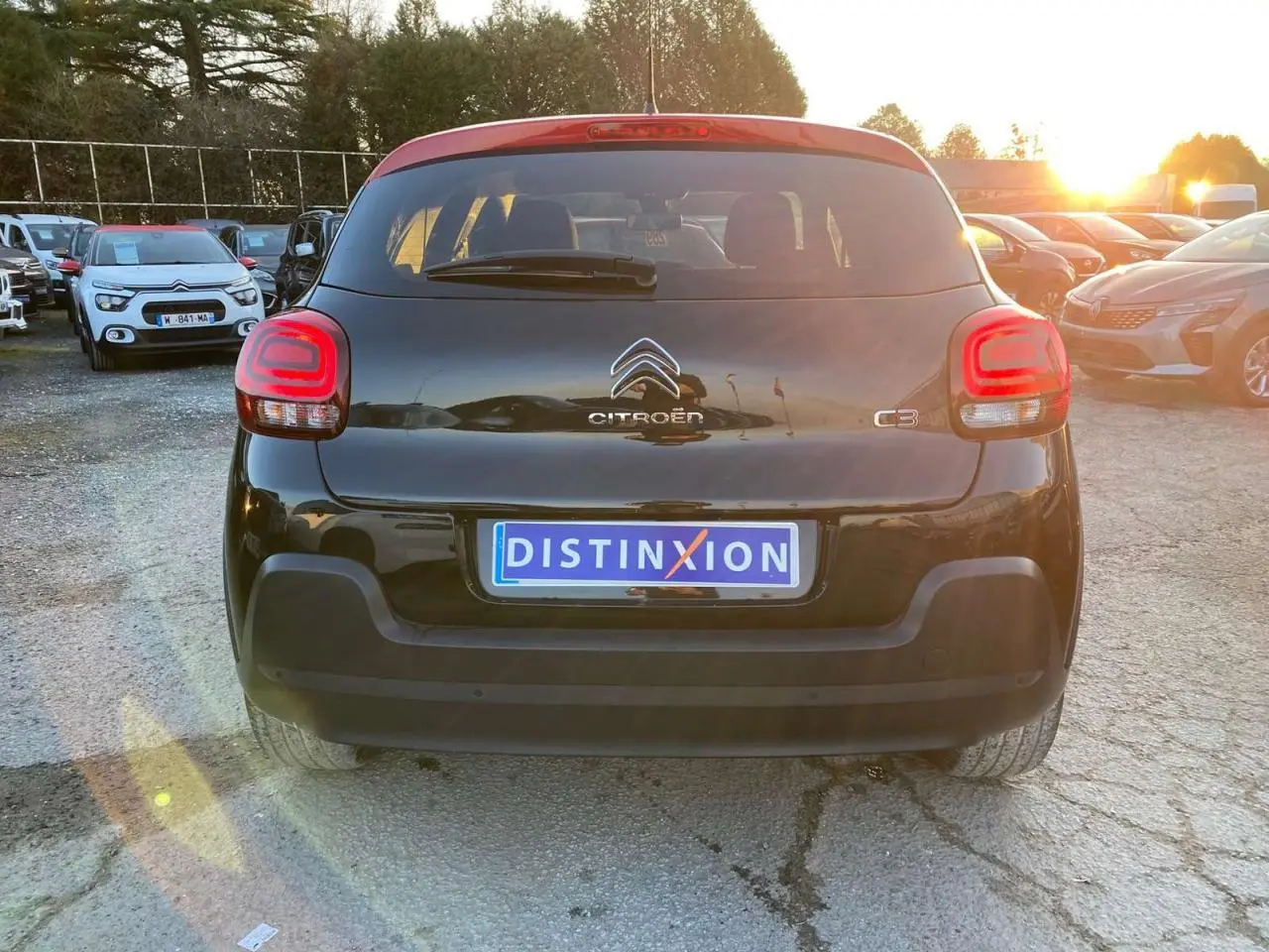 Vue arrière d'une Citroën C3 noire avec toit rouge, feux arrière allumés, stationnée en extérieur au coucher du soleil.
