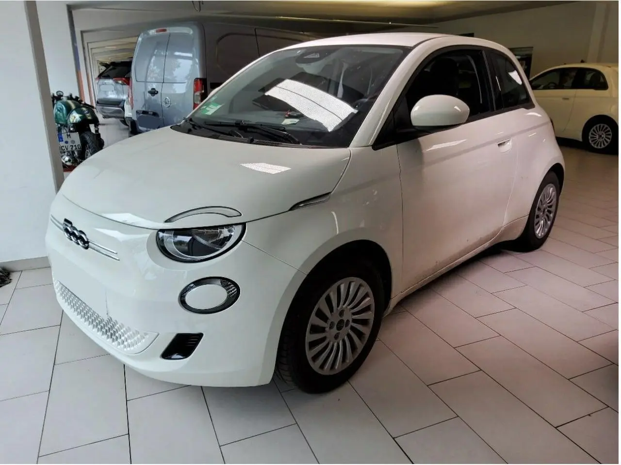 Vue 3/4 avant droite d'une Fiat 500 électrique blanche stationnée en intérieur avec jantes spécifiques et phares ronds LED.