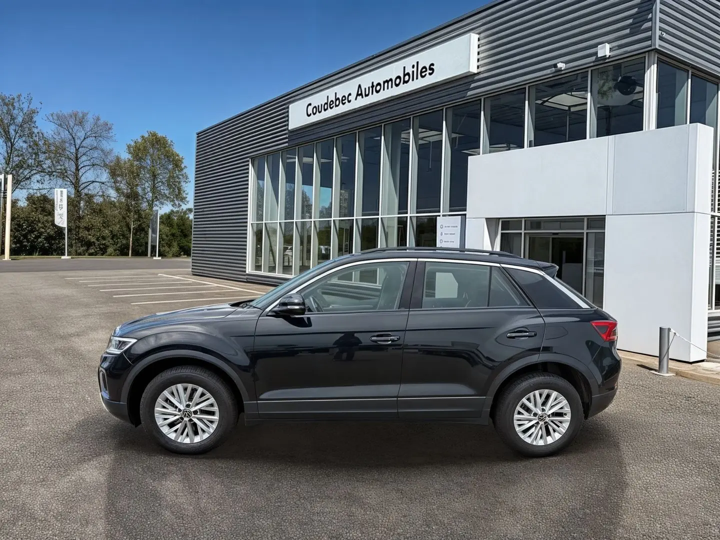 Vue de profil côté gauche d’un Volkswagen T-Roc noir 2025 garé devant un bâtiment de concession automobile.
