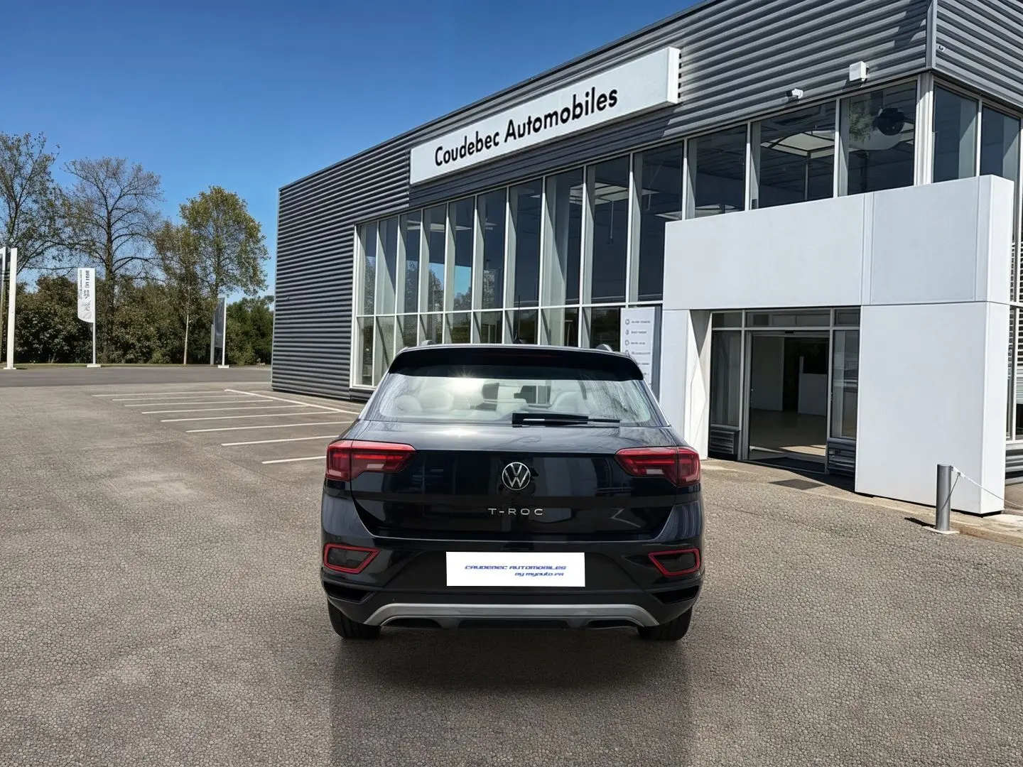 Vue arrière d’un Volkswagen T-Roc noir 2025 stationné devant un bâtiment moderne de concession automobile.