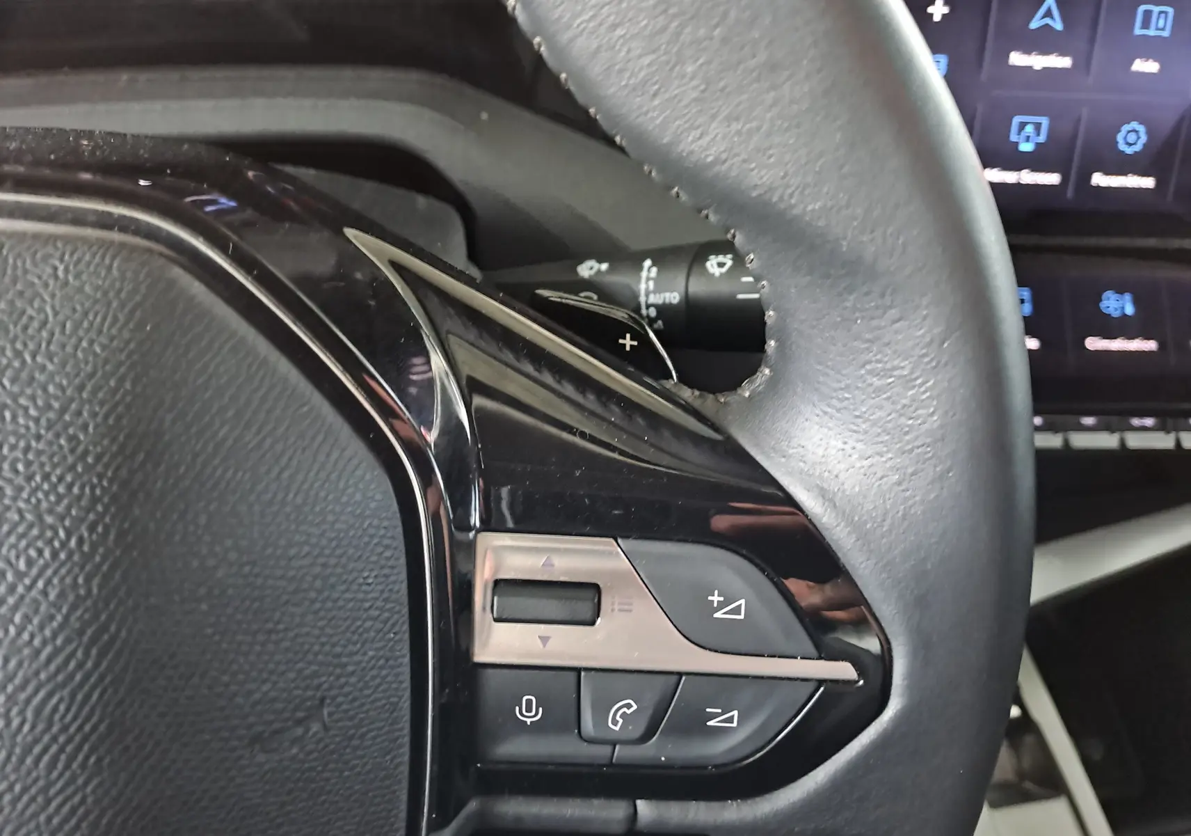 Gros plan sur les commandes du volant cuir noir de la Peugeot 308 1.5 BlueHDi 130ch Allure 2024 avec écran tactile en arrière-plan.