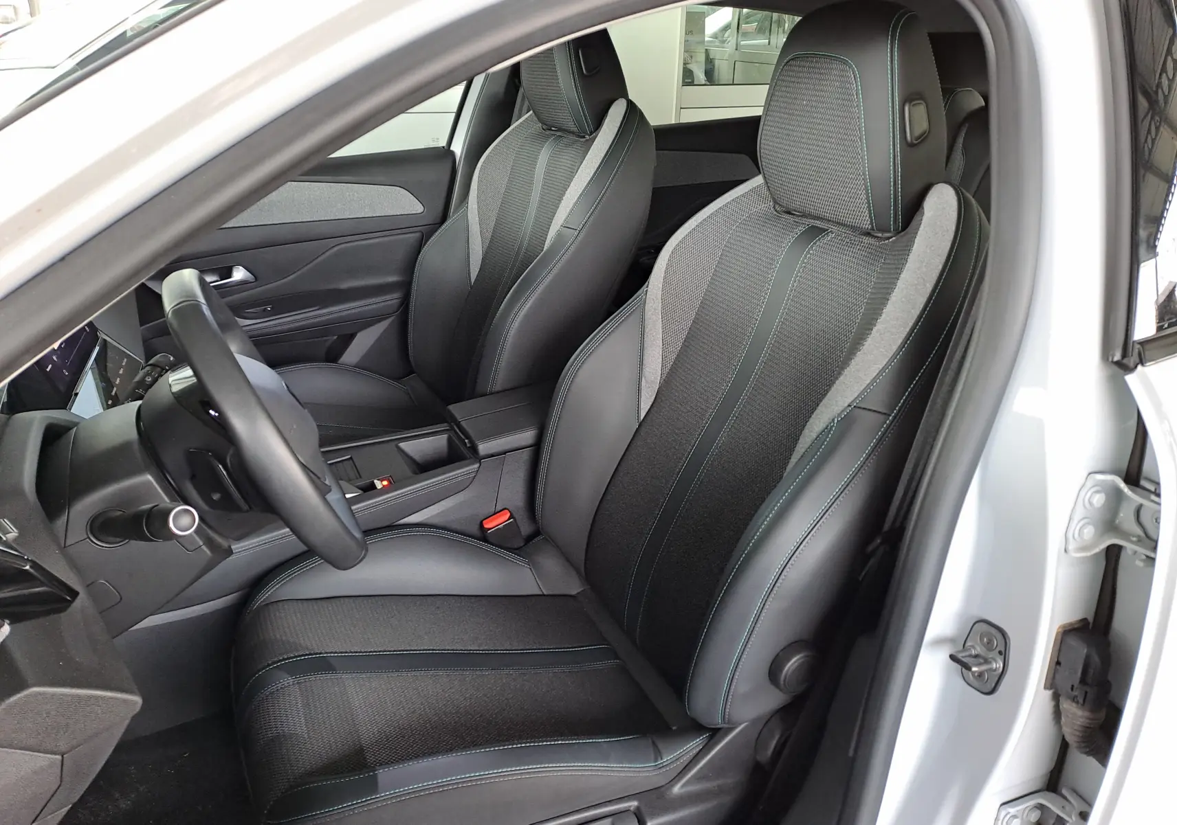 Vue intérieure côté conducteur de la Peugeot 308 2024 blanc Okenite, sièges noirs avec surpiqûres bleues et tissu mixte.