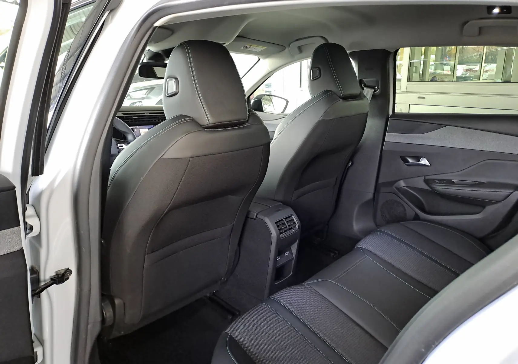 Vue intérieure arrière droite d'une Peugeot 308 blanche Okenite, sièges noirs en tissu et cuir avec aérateurs centraux visibles.
