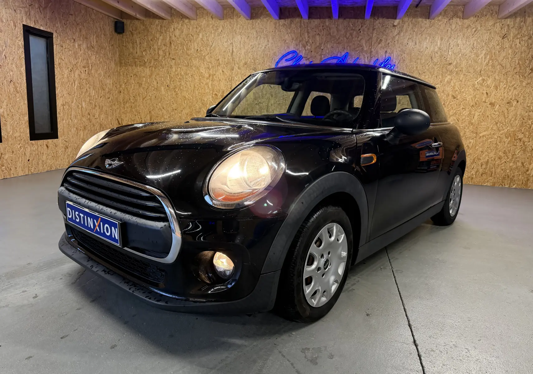 Mini One noire vue 3/4 avant droit en intérieur, phares et antibrouillards allumés, calandre chromée visible.