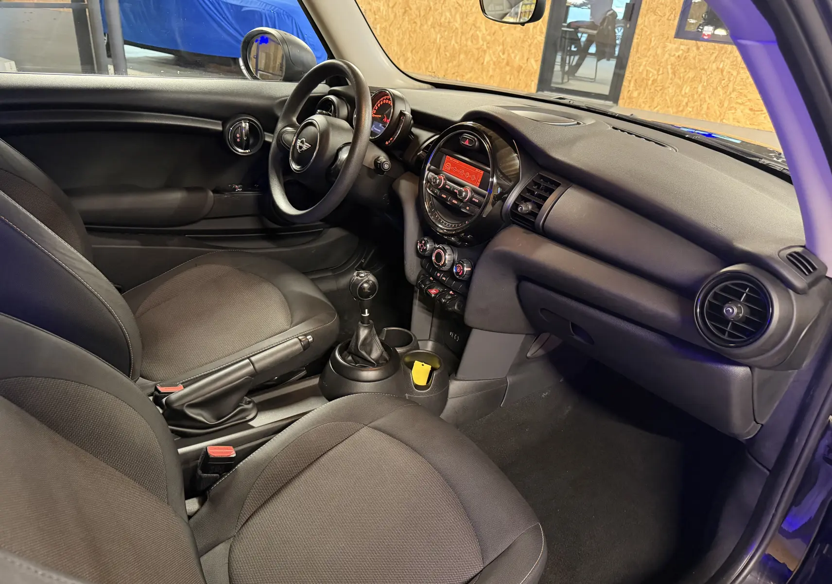 Vue intérieure côté conducteur de la MINI 1.5i noire, avec tableau de bord rond et levier de vitesses manuel visible.