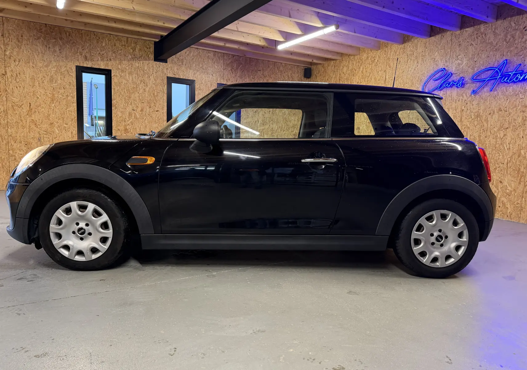 Vue de profil côté gauche d'une MINI noire 3 portes, stationnée en intérieur avec mur en bois et éclairage bleu.