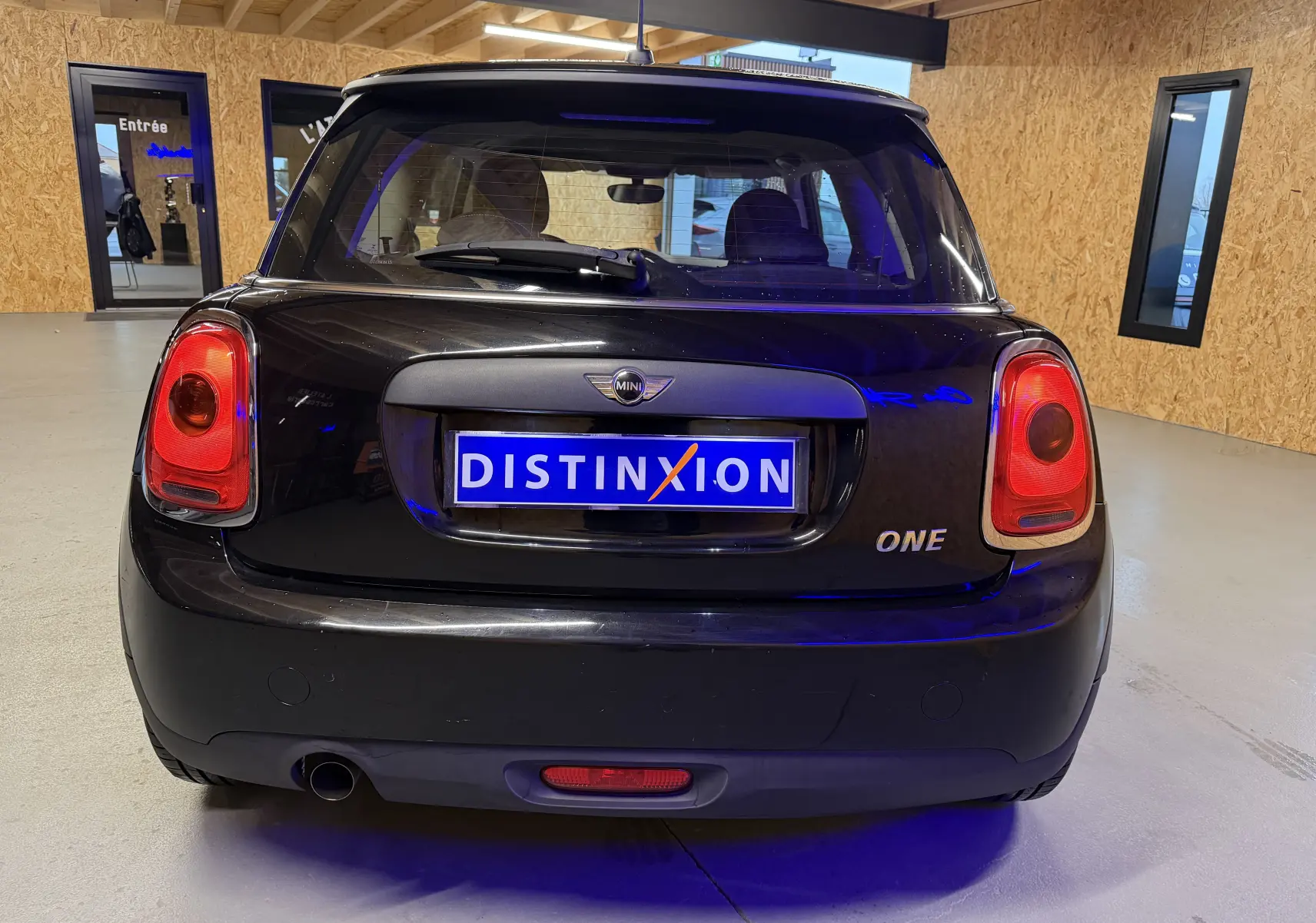 Vue arrière d'une MINI One noire de 2018, avec feux rouges et logo MINI bien visibles dans un intérieur en bois.