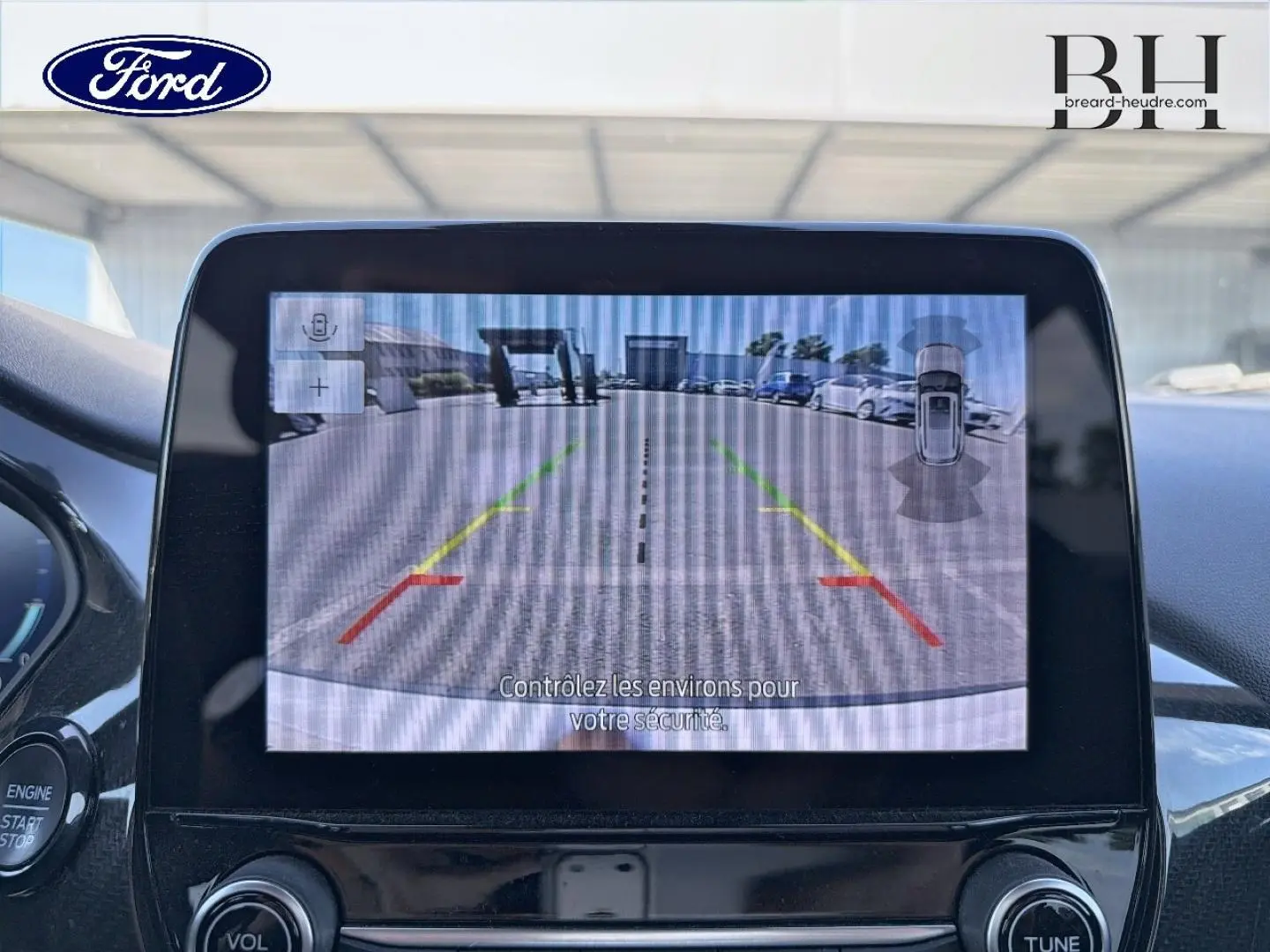 Écran tactile intérieur montrant la caméra de recul avec vue arrière sur un parking et véhicules stationnés.