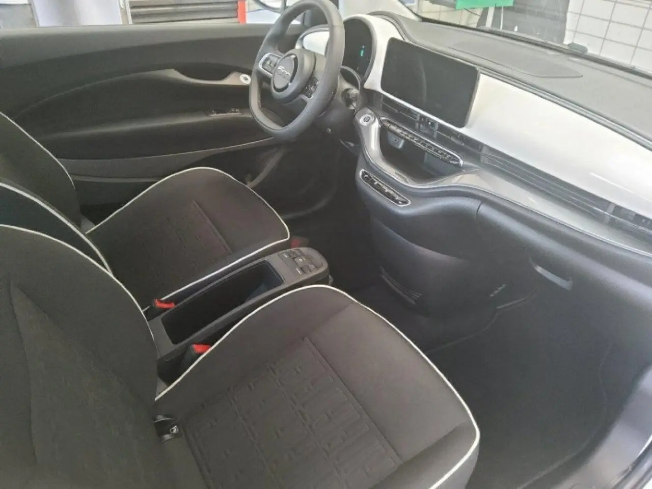 Vue intérieure côté conducteur de la Fiat 500 électrique 2024, sièges noirs avec surpiqûres blanches et tableau de bord blanc.