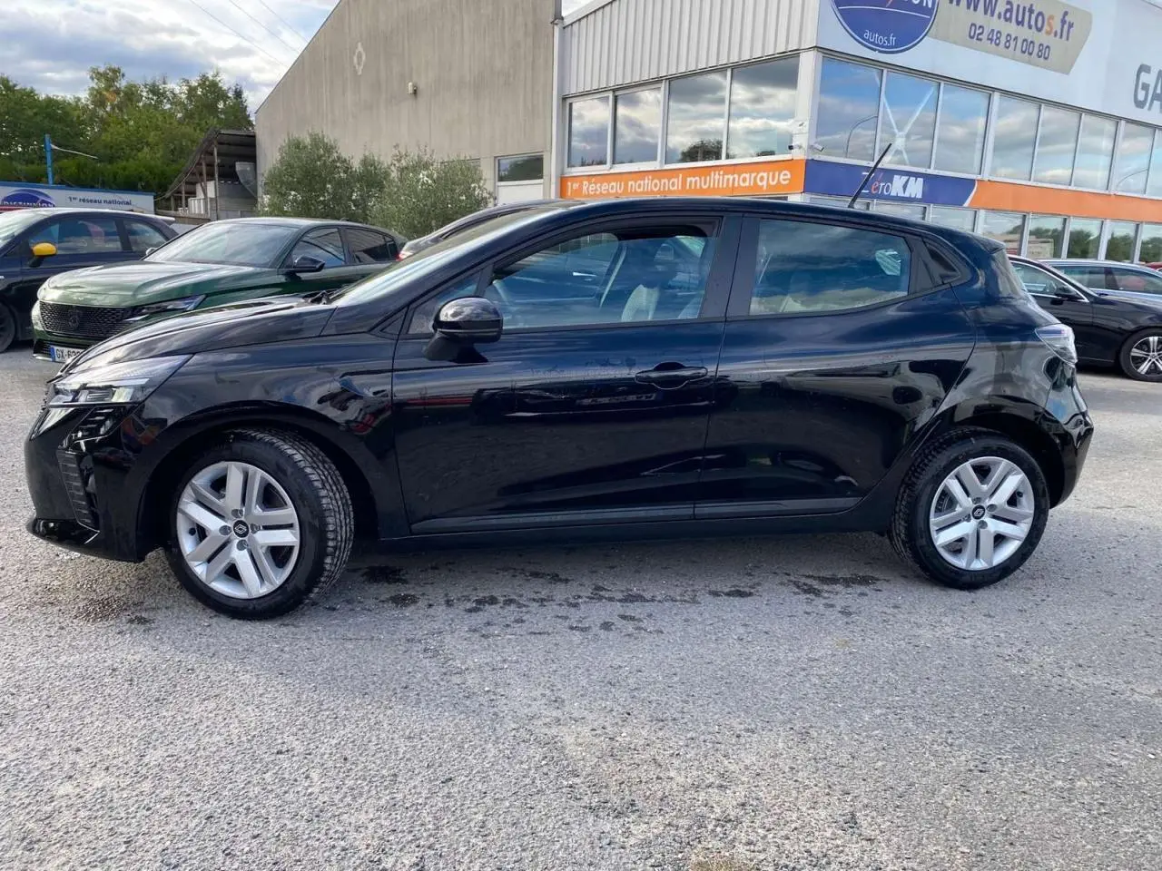 Profil côté gauche d'une Renault Clio 2025 noire étoile, garée devant un bâtiment commercial avec d'autres voitures visibles.