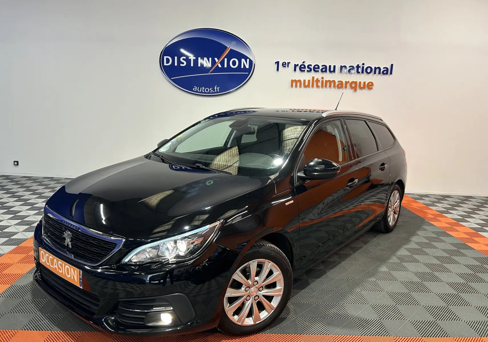 Peugeot 308 SW noire vue en 3/4 avant droit, avec jantes alliage et feux allumés dans un showroom.