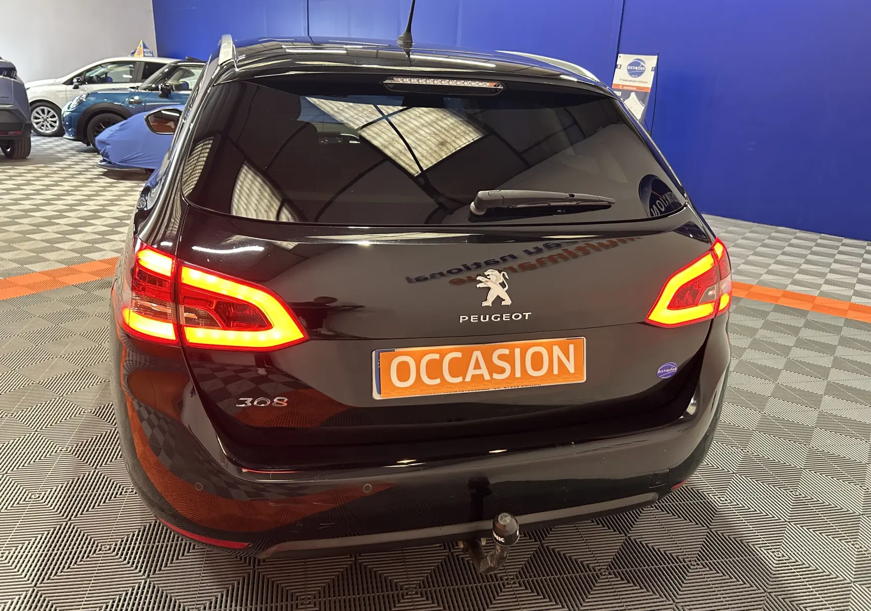 Vue 3/4 arrière d'une Peugeot 308 SW noire avec feux allumés et attelage visible en intérieur showroom.