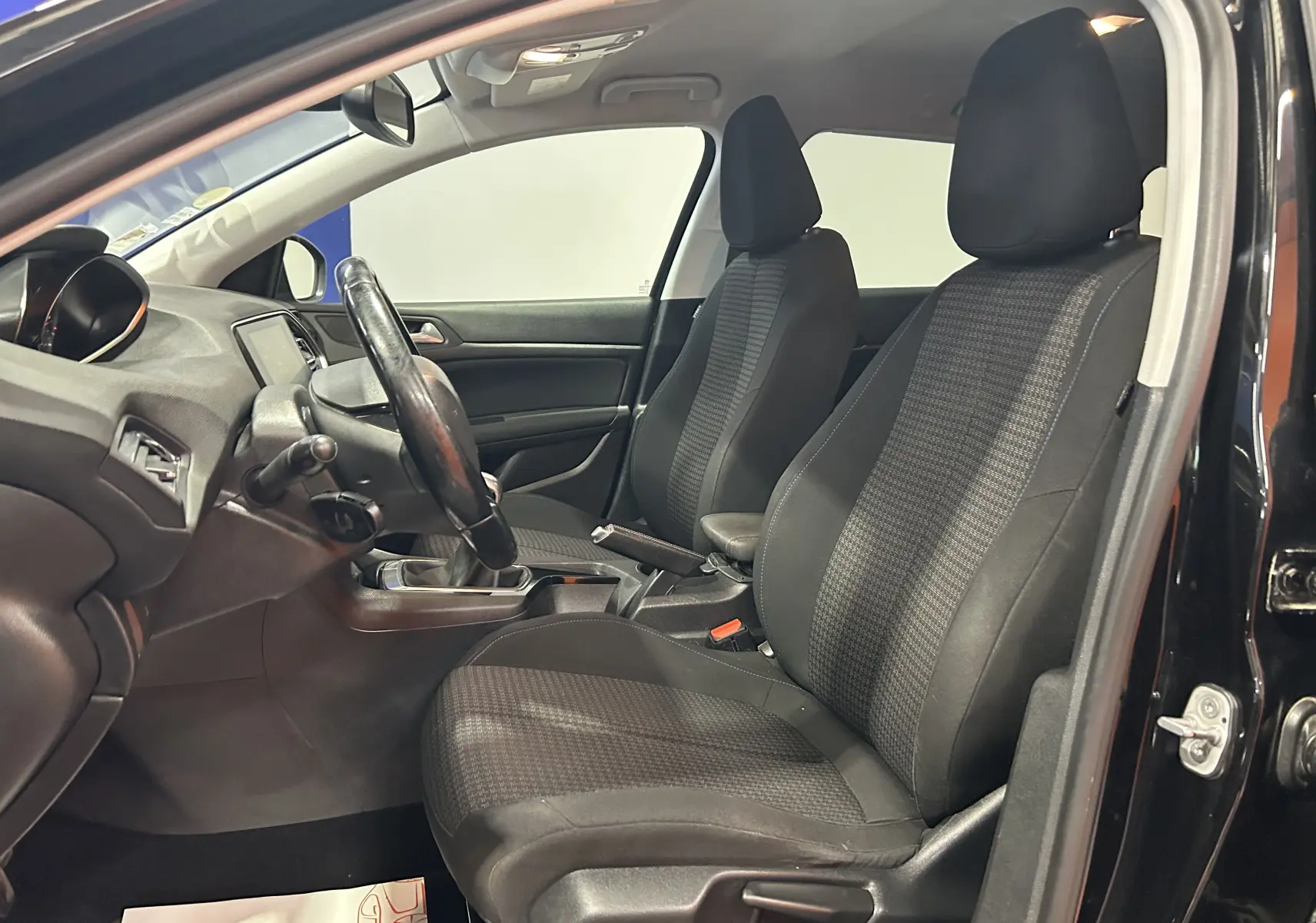 Vue intérieure côté conducteur de la Peugeot 308 SW noire, montrant les sièges tissu et le volant cuir.