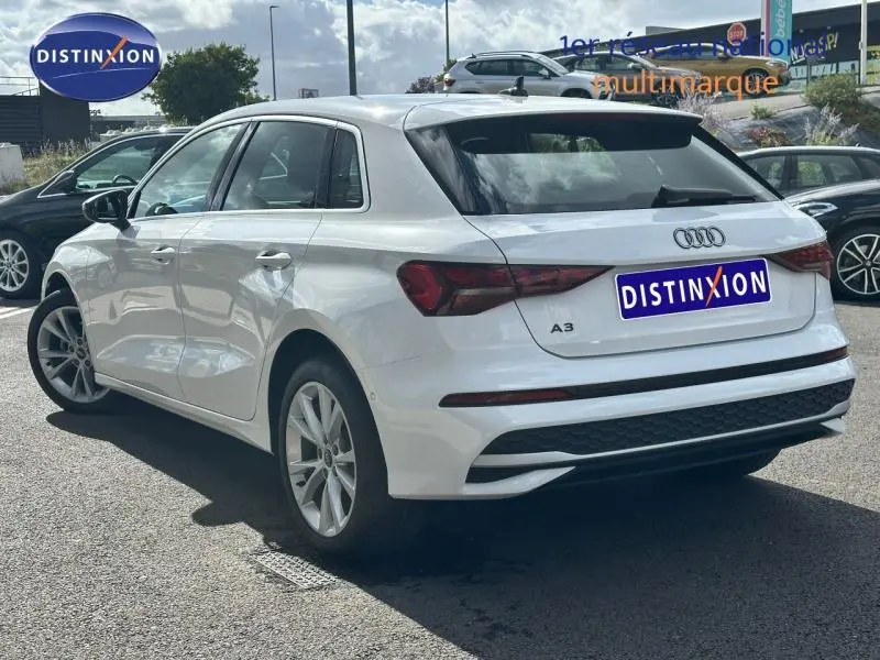 Audi A3 Sportback blanche vue 3/4 arrière droit, avec feux arrière LED et becquet noir sur parking extérieur