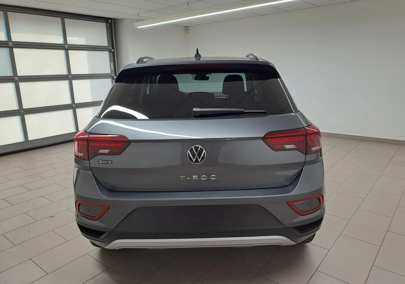 Vue arrière d'un Volkswagen T-Roc gris Indium 2024 avec feux LED et caméra de recul dans un garage lumineux.