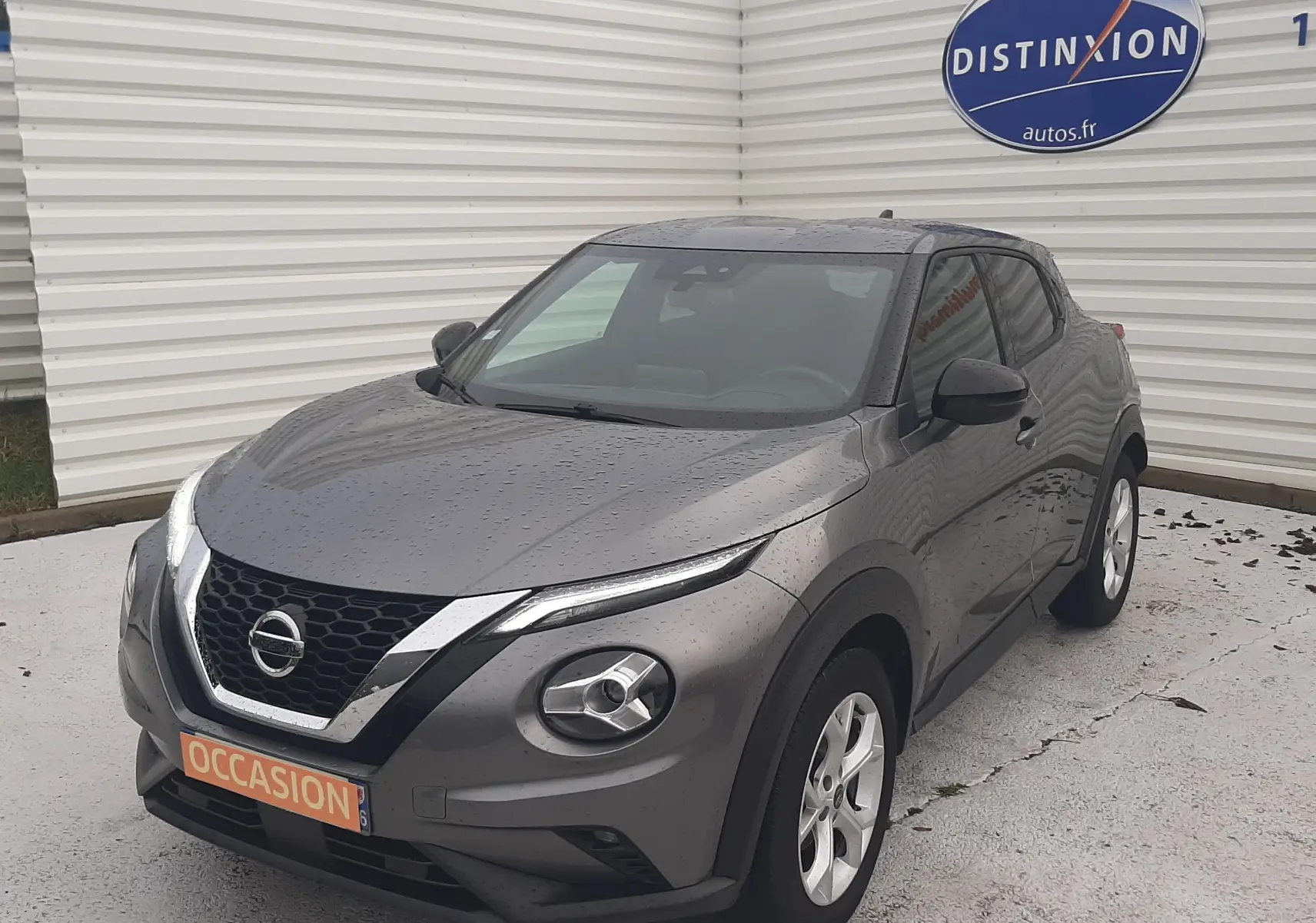 Nissan Juke gris 3/4 avant droit sous la pluie, avec calandre noire et phares ronds distinctifs.