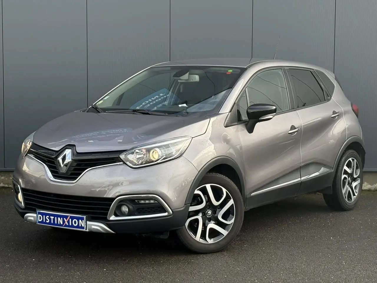 Renault Captur gris Cassiopée en 3/4 avant droit, avec jantes alliage noires et phares allumés.