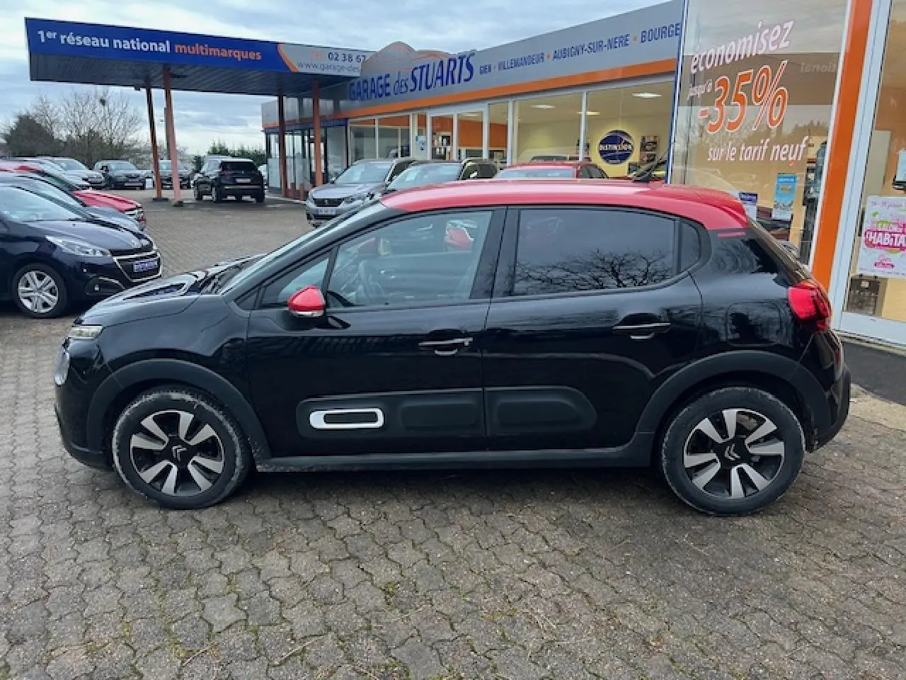 Profil latéral gauche d'une Citroën C3 Shine 2023 noire avec toit rouge et jantes alliage diamantées, stationnée devant un garage.