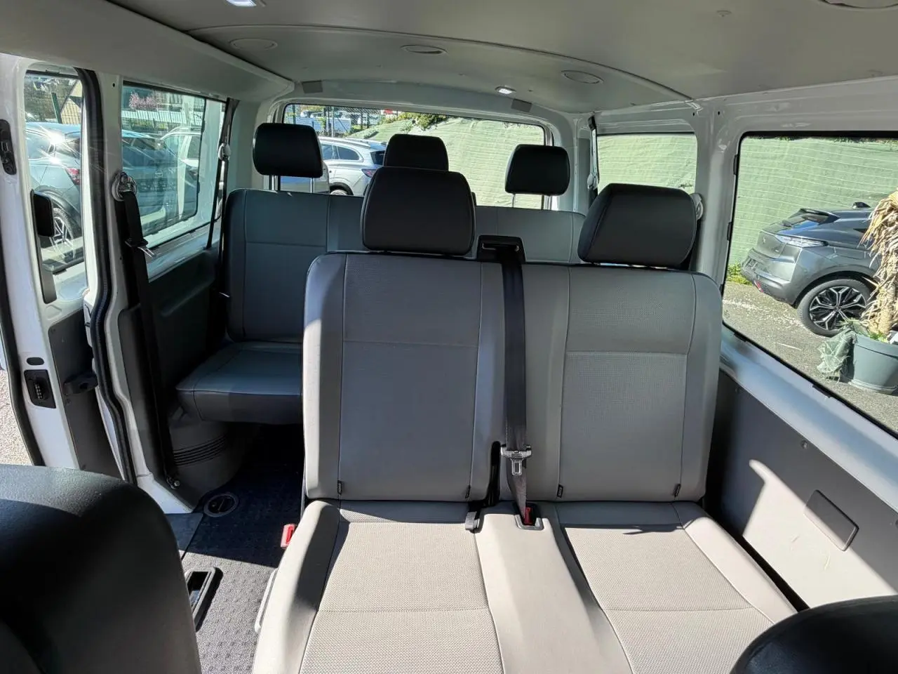 Vue intérieure du Volkswagen Transporter T6 Minibus blanc, montrant les sièges arrière en tissu gris et la porte coulissante ouverte côté gauche.
