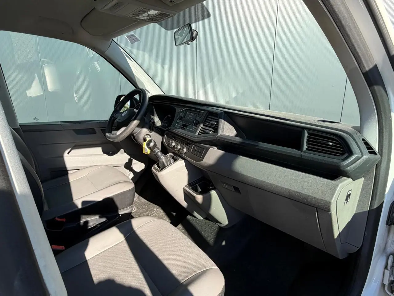 Vue intérieure côté conducteur du Volkswagen Transporter T6 blanc, montrant le tableau de bord et les sièges avant gris.