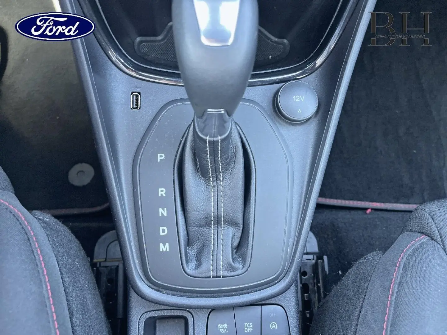 Gros plan sur la console centrale de la Ford Puma 2022, boîte auto avec levier cuir et coutures blanches, sièges tissu noir à surpiqûres rouges.