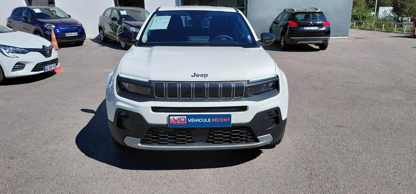 Vue avant d'un Jeep Avenger blanc 2025 avec calandre noire et plaque "Véhicule récent" sur un parking extérieur.