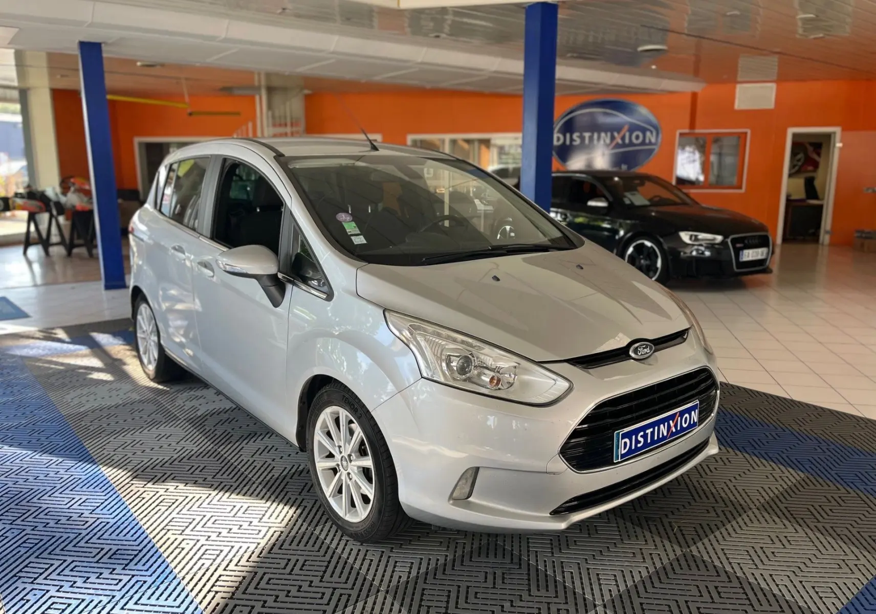 Ford B-MAX gris clair en 3/4 avant droit dans un showroom avec plaque Distinxion visible