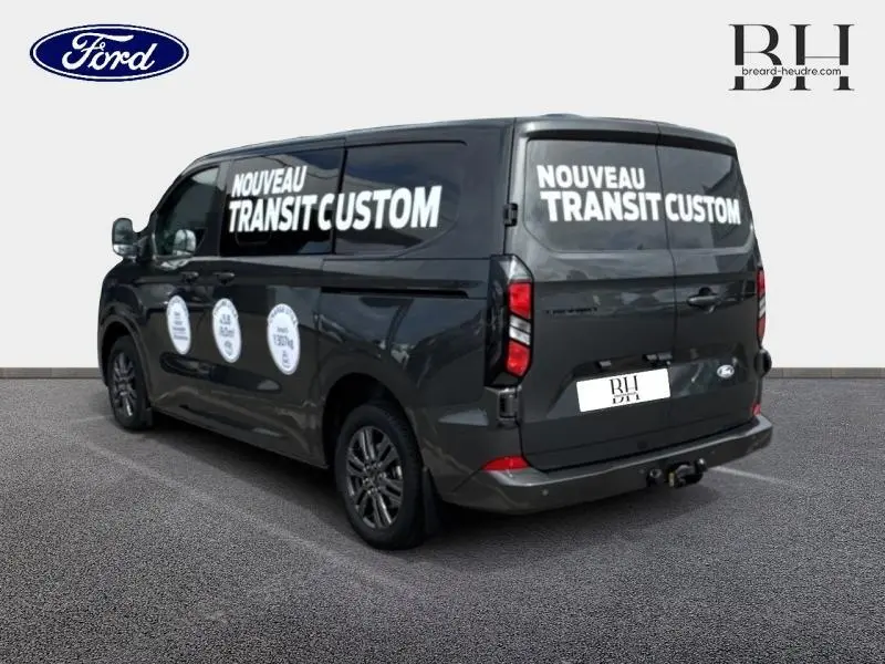Vue 3/4 arrière droite du Ford Transit Custom gris métallisé avec marquages publicitaires blancs sur les vitres latérales et arrière.