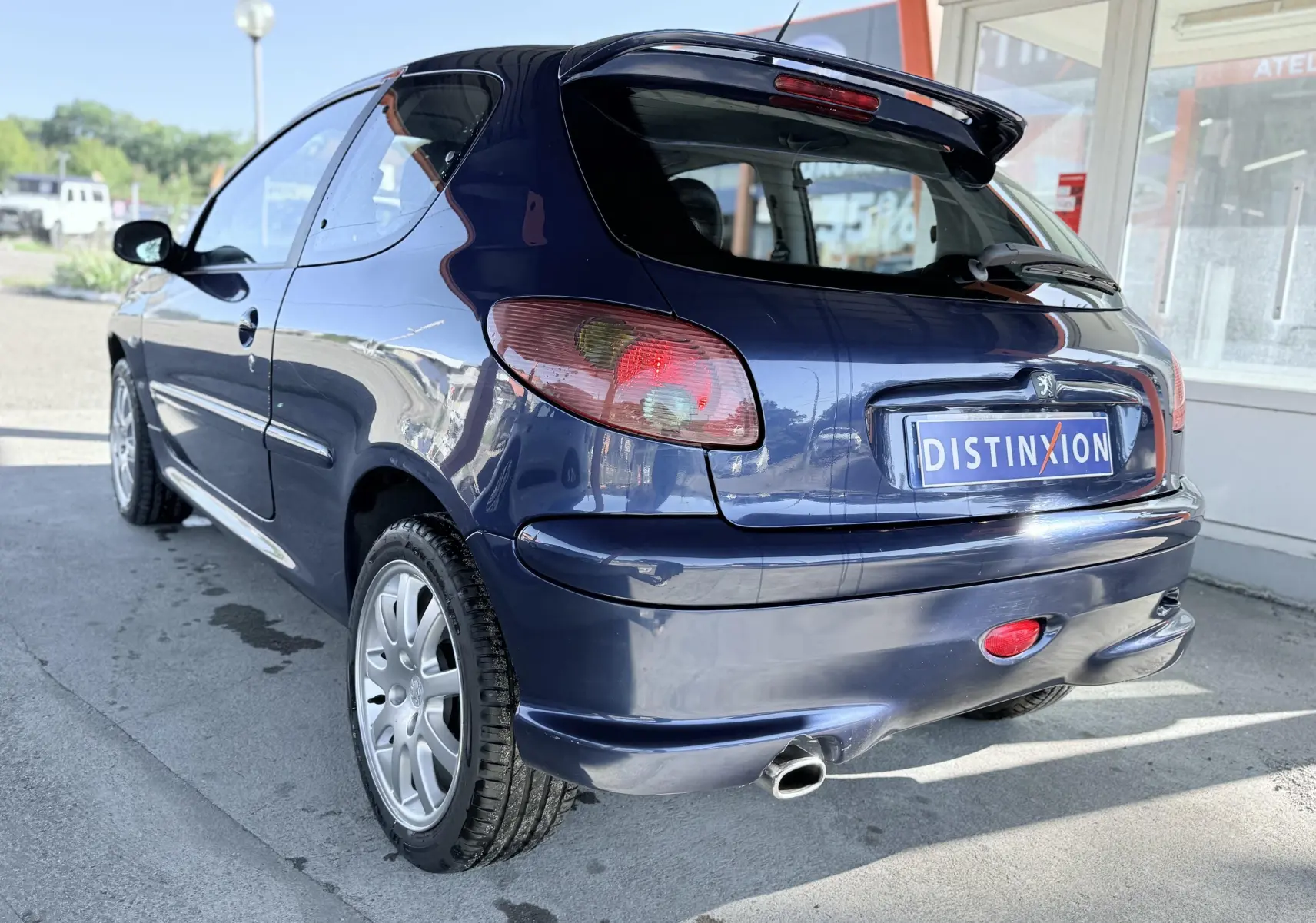 Peugeot 206 bleu vue 3/4 arrière droit, avec hayon ouvert et jantes alliage visibles.
