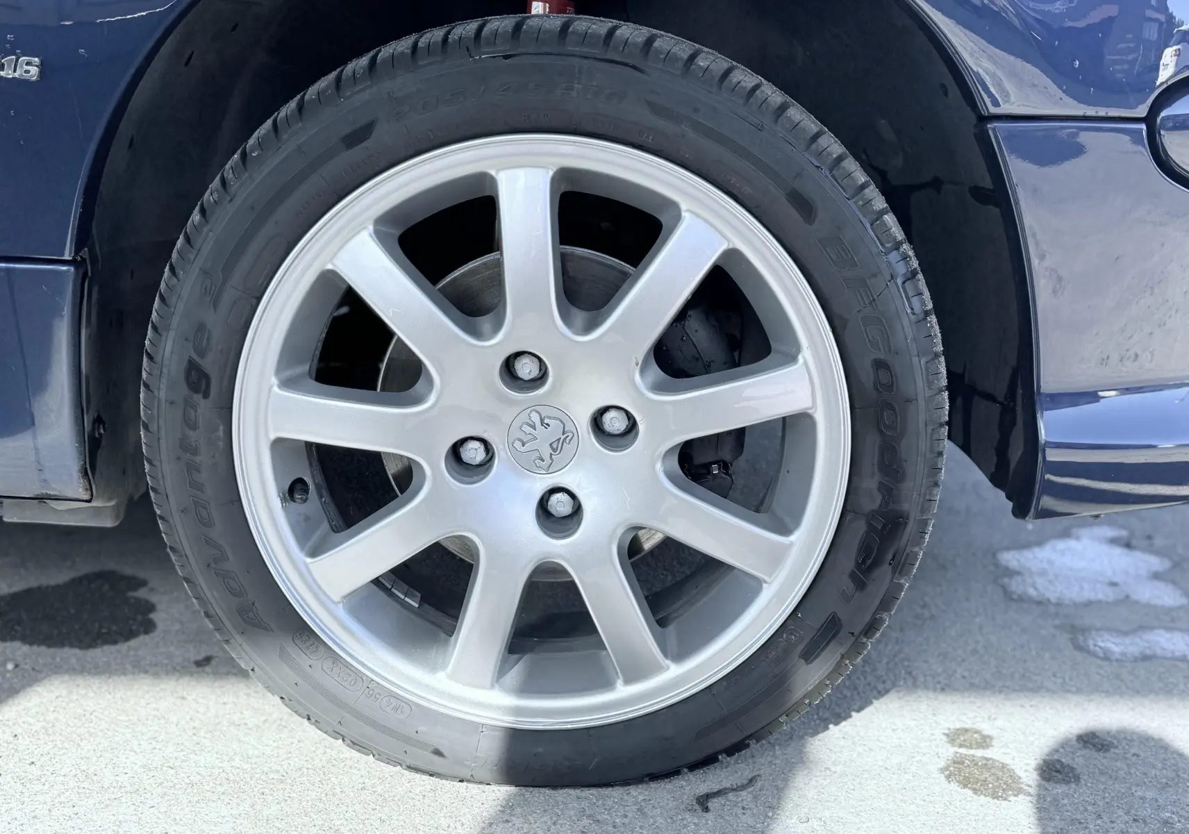 Gros plan sur la roue avant gauche d’une Peugeot 206 bleu 2003 avec jante alliage argentée et logo lion au centre.