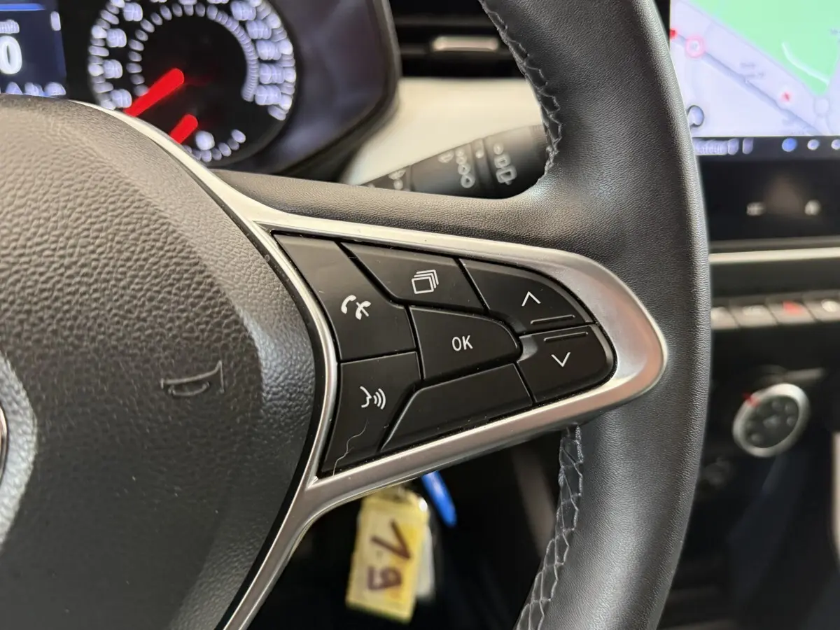 Gros plan sur les commandes au volant cuir noir de la Renault Clio blanche, avec tableau de bord et écran tactile en arrière-plan.