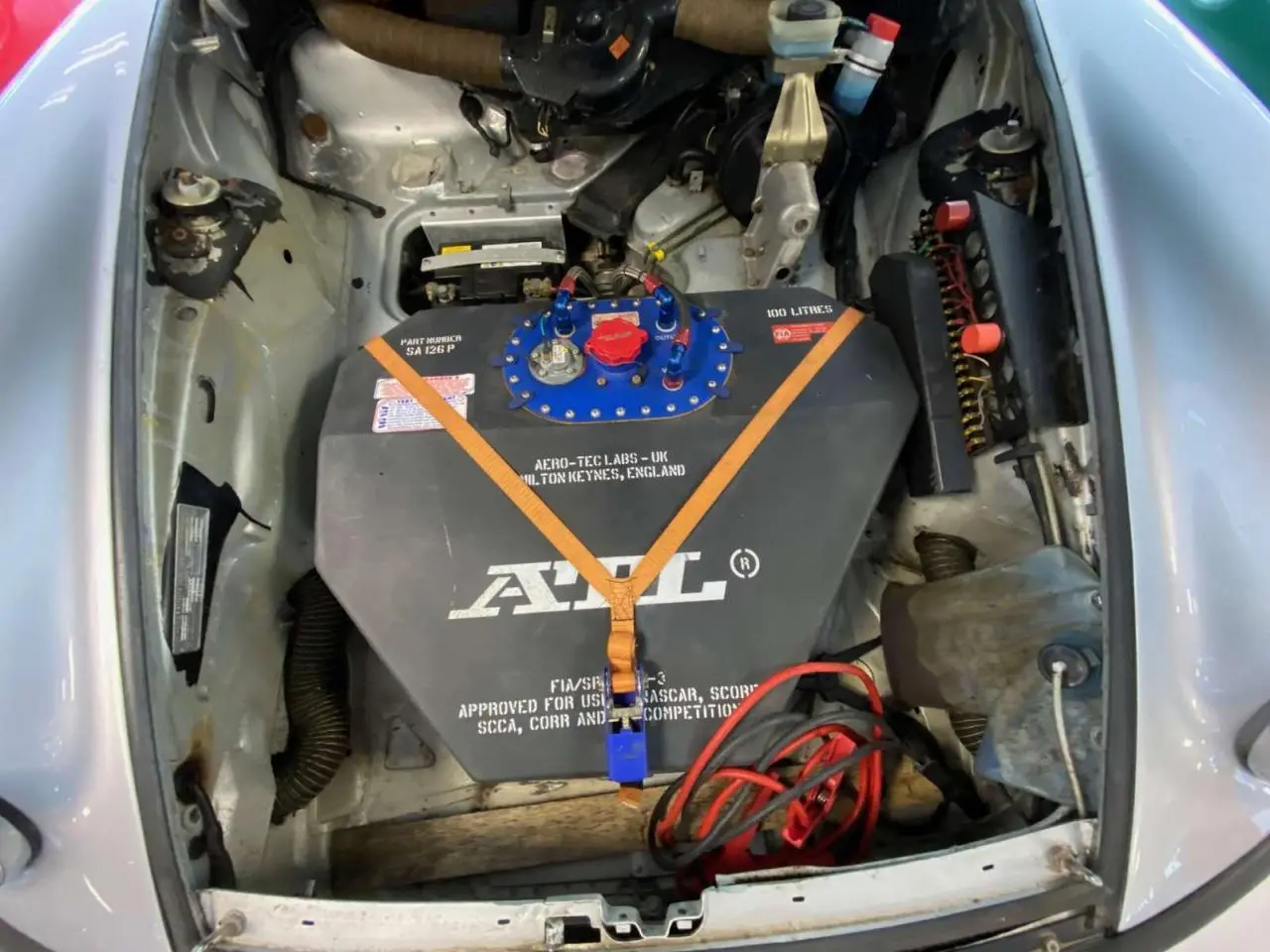 Vue plongeante sur le compartiment avant gris métallisé d’une Porsche 911 3.0 SC avec réservoir FIA et sangles orange.