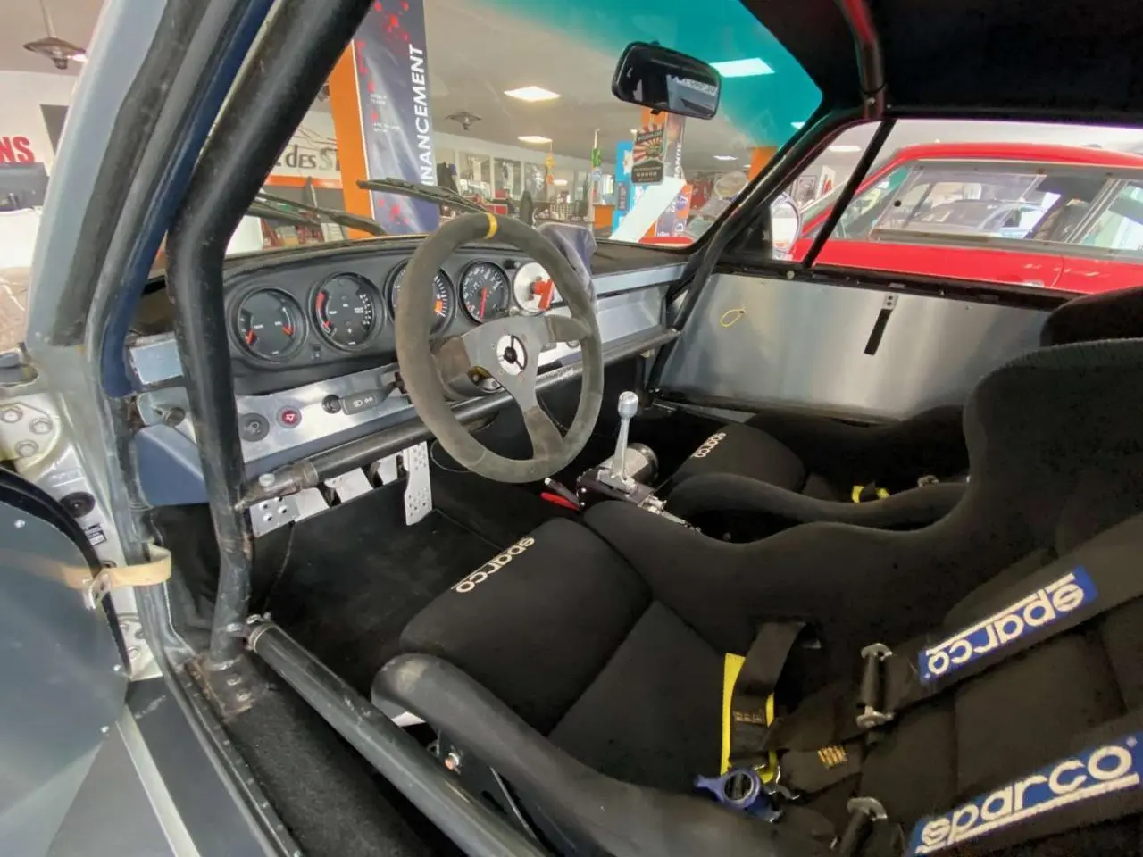 Vue intérieure côté conducteur de la Porsche 911 3.0 SC gris métallisé, avec baquets Sparco et arceau de sécurité.