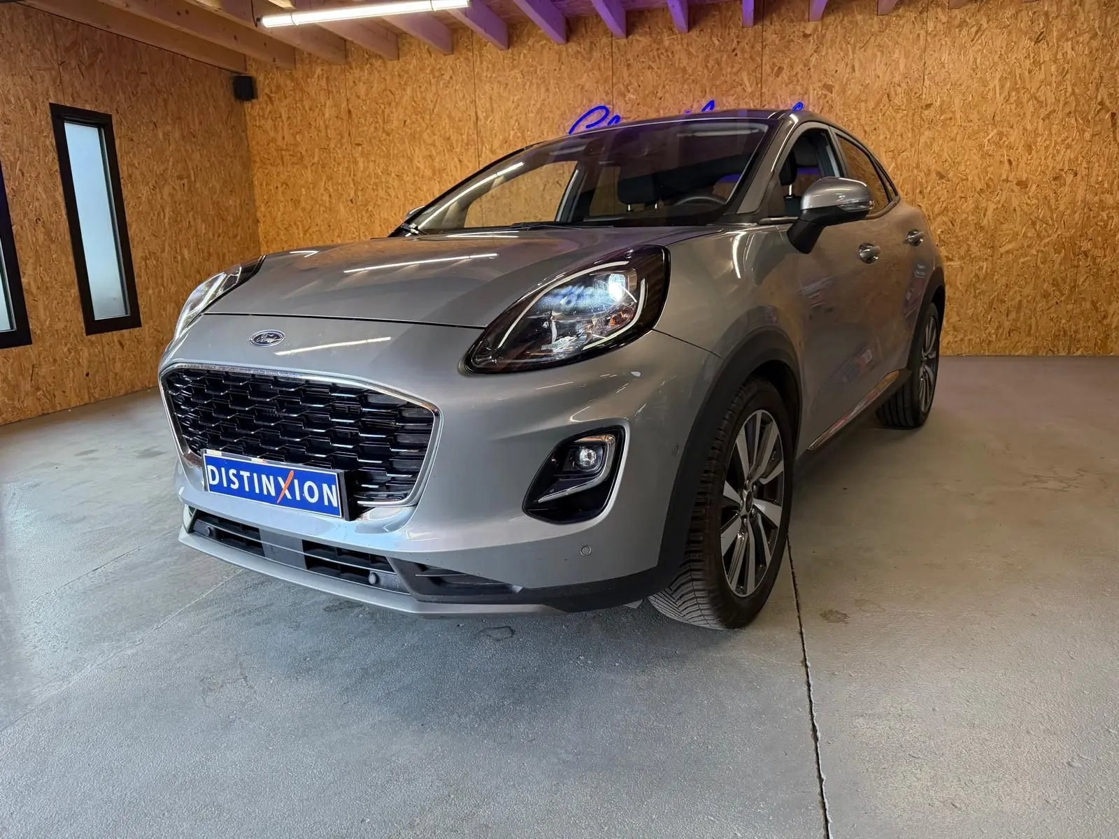Ford Puma gris en 3/4 avant droit, avec calandre noire et phares allumés dans un garage en bois.