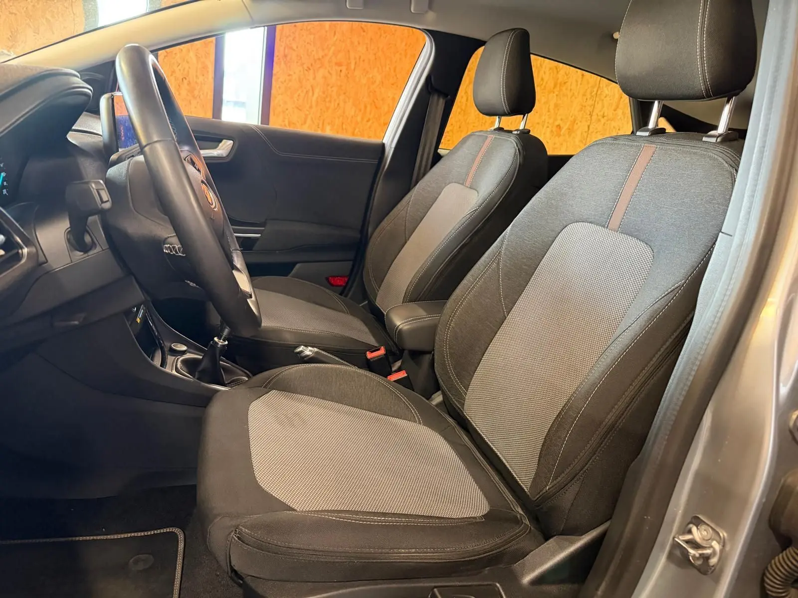 Vue intérieure côté gauche du Ford Puma gris 2021, montrant les sièges avant en tissu noir et gris avec volant et levier de vitesses manuelle.