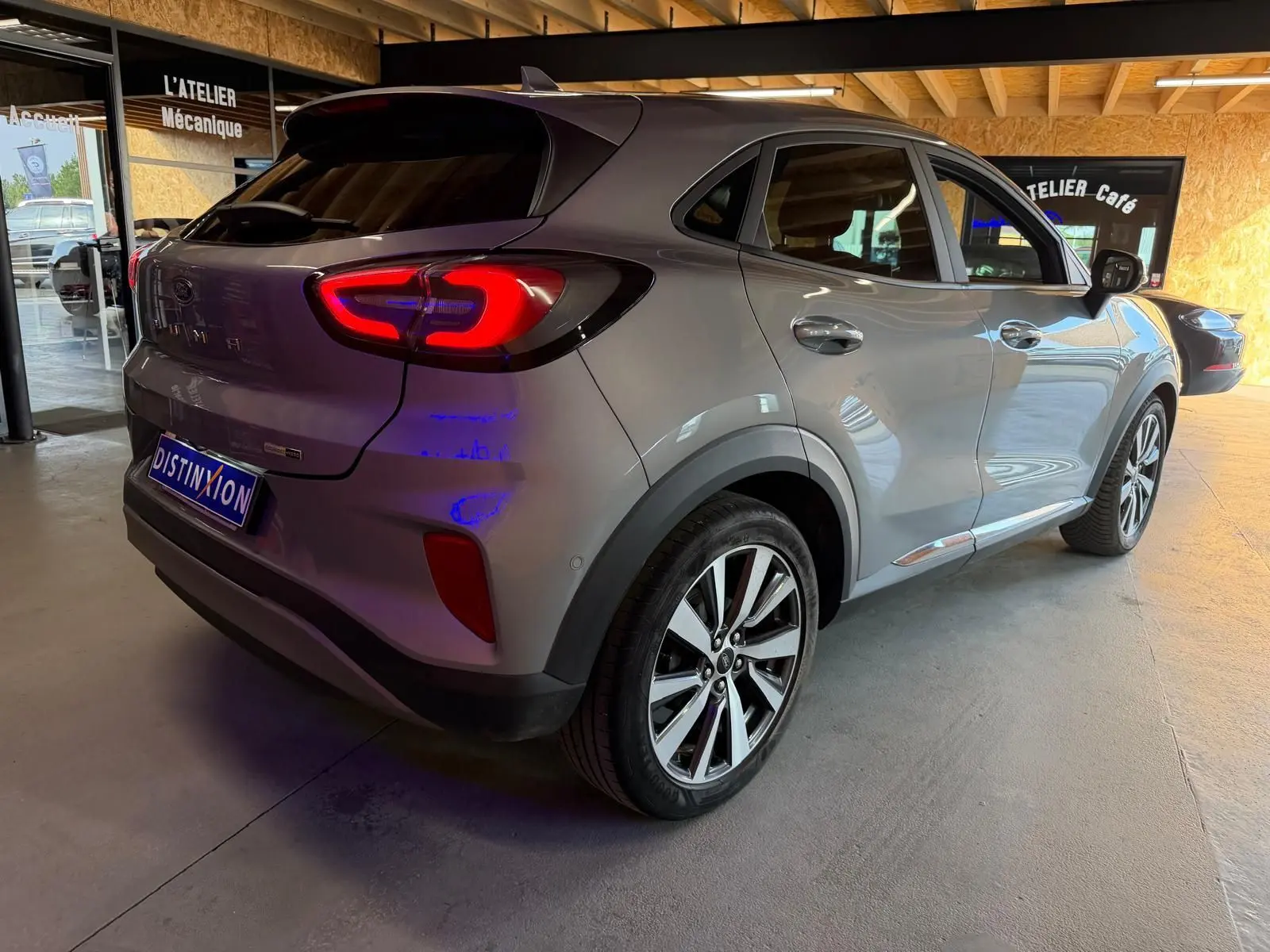 Vue 3/4 arrière droite d'un Ford Puma gris 2021 avec feux arrière allumés dans un garage lumineux.