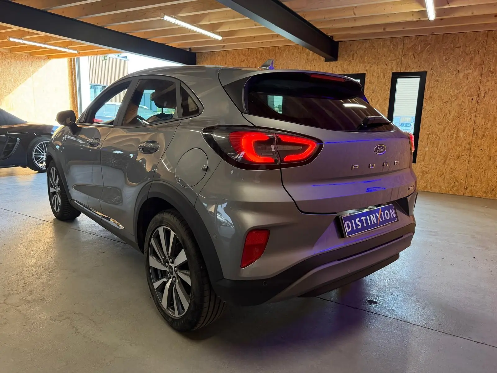Vue 3/4 arrière droite d'une Ford Puma gris métallisé 2021 avec feux arrière allumés dans un garage lumineux.