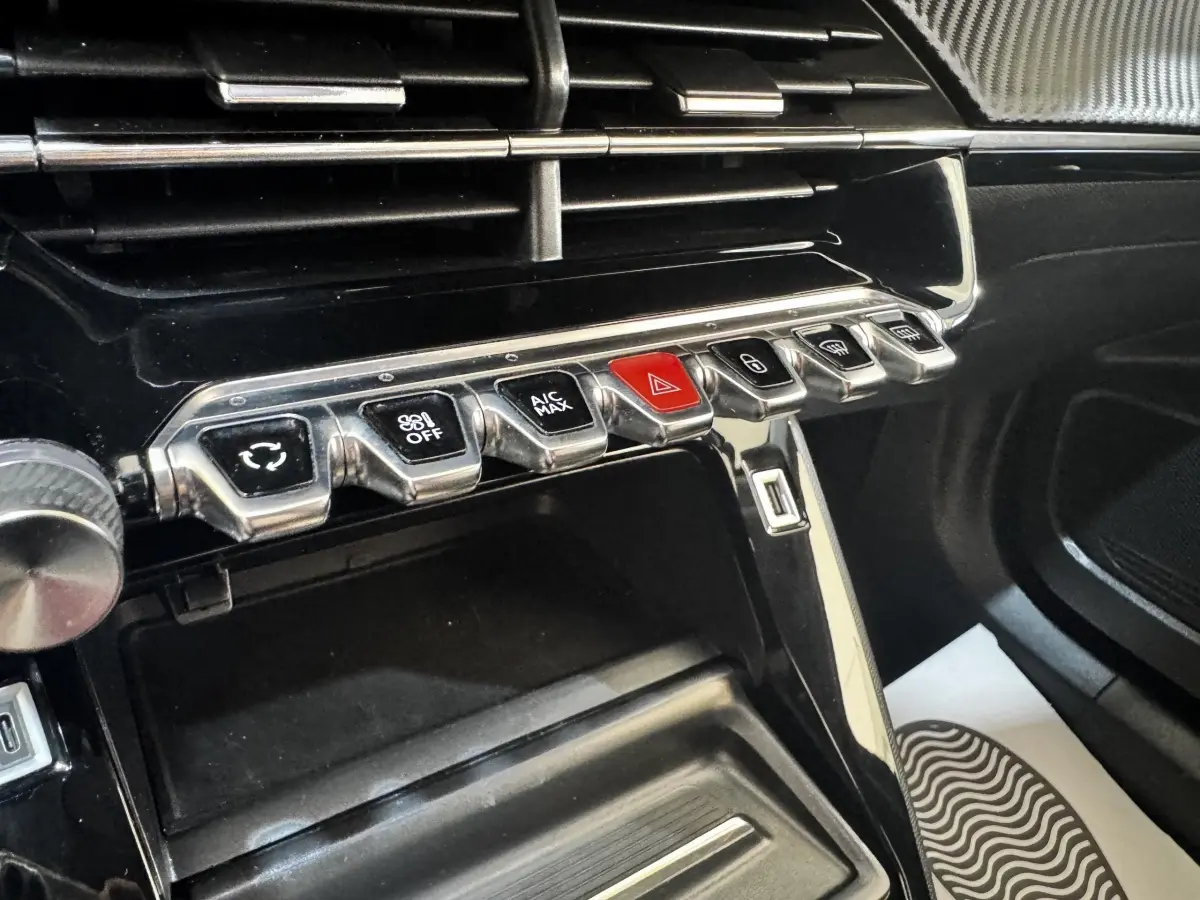 Gros plan sur la console centrale noire du Peugeot 2008 2022, avec rangements et rangée de boutons chromés dont le bouton d'alerte rouge.