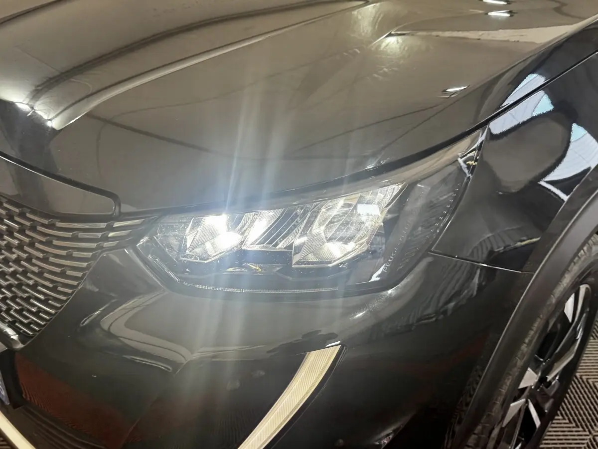Gros plan sur le phare avant droit allumé d’un Peugeot 2008 noir, mettant en valeur la calandre et la carrosserie brillante.
