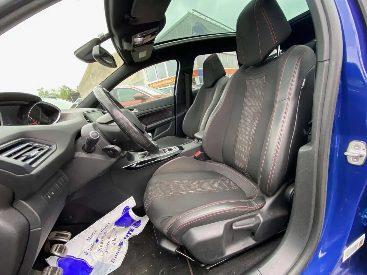 Vue intérieure côté conducteur de la Peugeot 308 SW bleu magnétique, sièges TEP/tissu noirs avec surpiqûres rouges et toit panoramique.