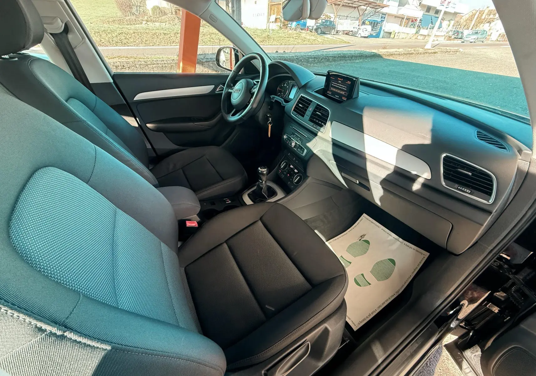 Vue intérieure côté conducteur de l'Audi Q3 noir 2016, montrant les sièges tissu noirs et le tableau de bord avec écran central.