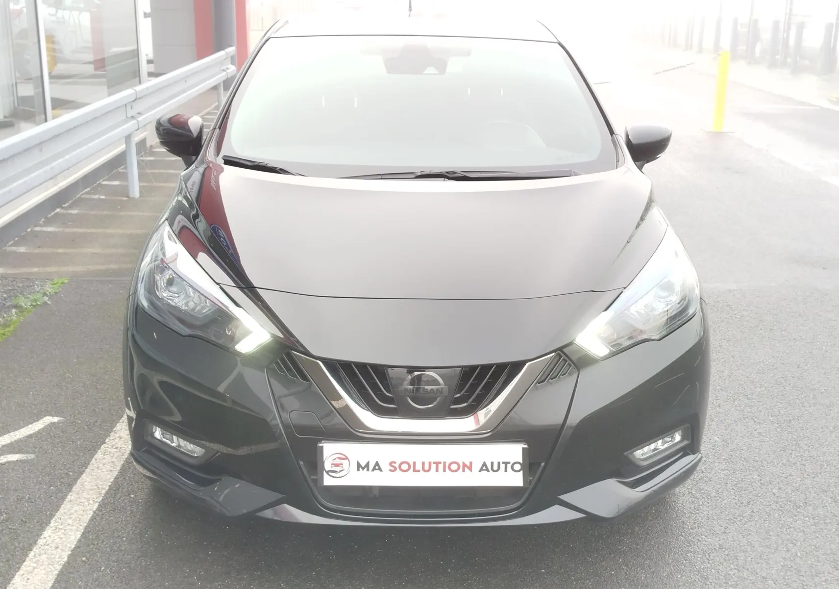 Vue frontale d'une Nissan Micra noire avec phares allumés et calandre chromée, stationnée sur un parking extérieur.