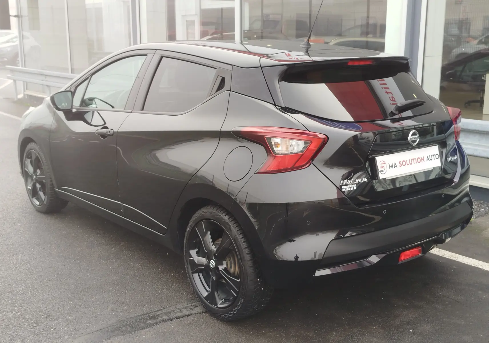 Vue 3/4 arrière droite d'une Nissan Micra noire avec jantes noires et vitres teintées, stationnée devant un showroom.