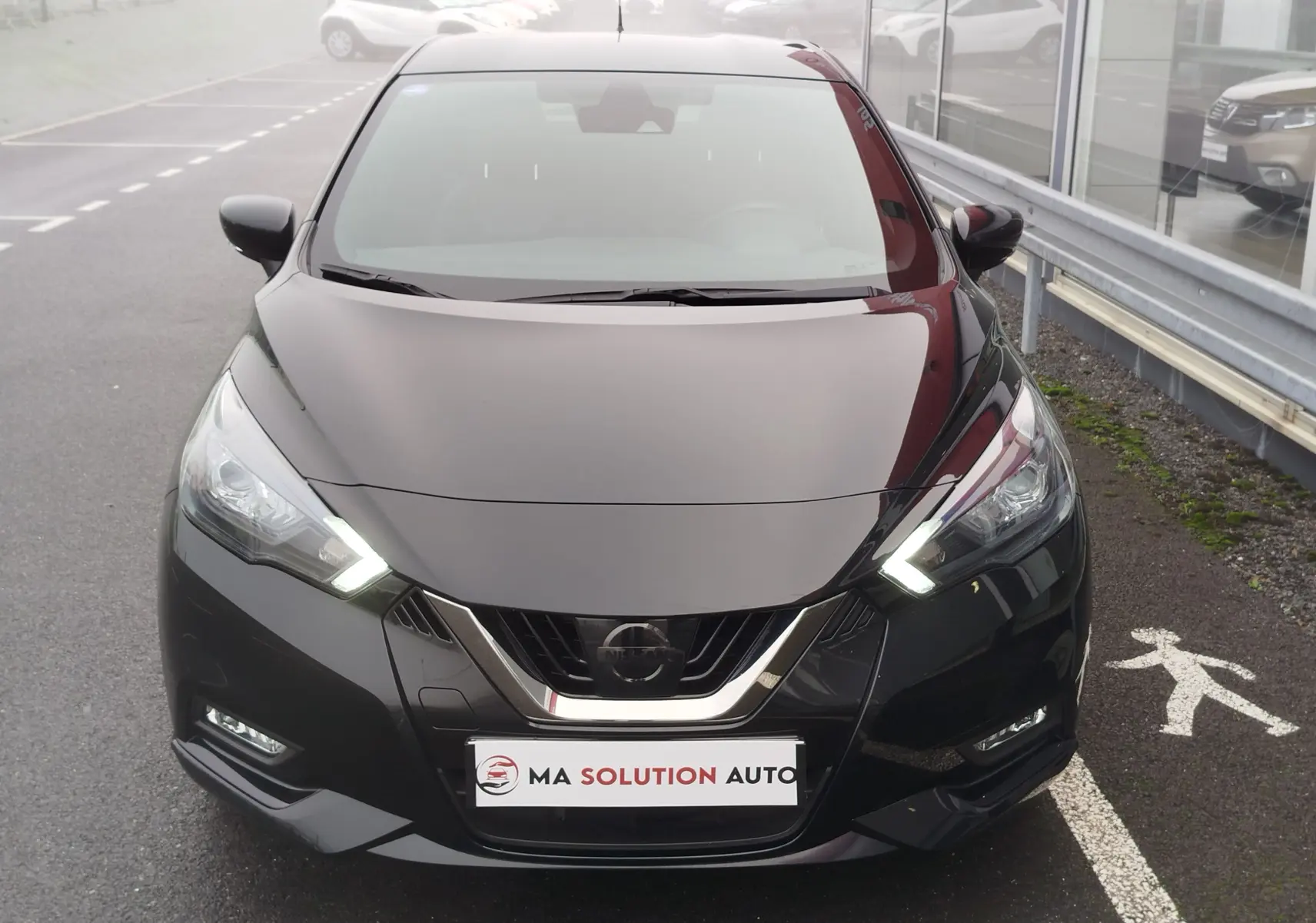 Vue frontale d'une Nissan Micra 1.0 IG-T 92ch N-Sport noire avec phares LED allumés sur parking extérieur.