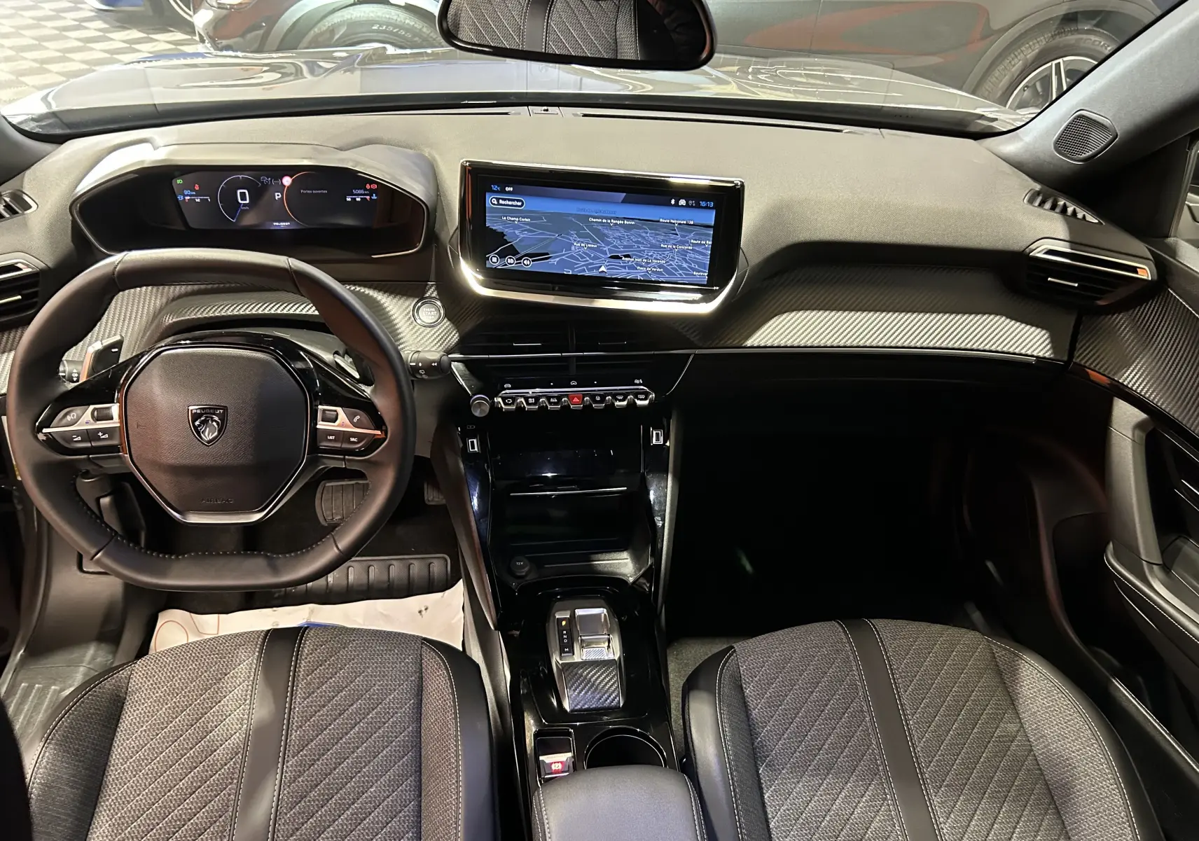 Vue intérieure frontale du tableau de bord et volant du Peugeot 2008 II Ph.2 Allure Hybrid 145 en finition noire et carbone.