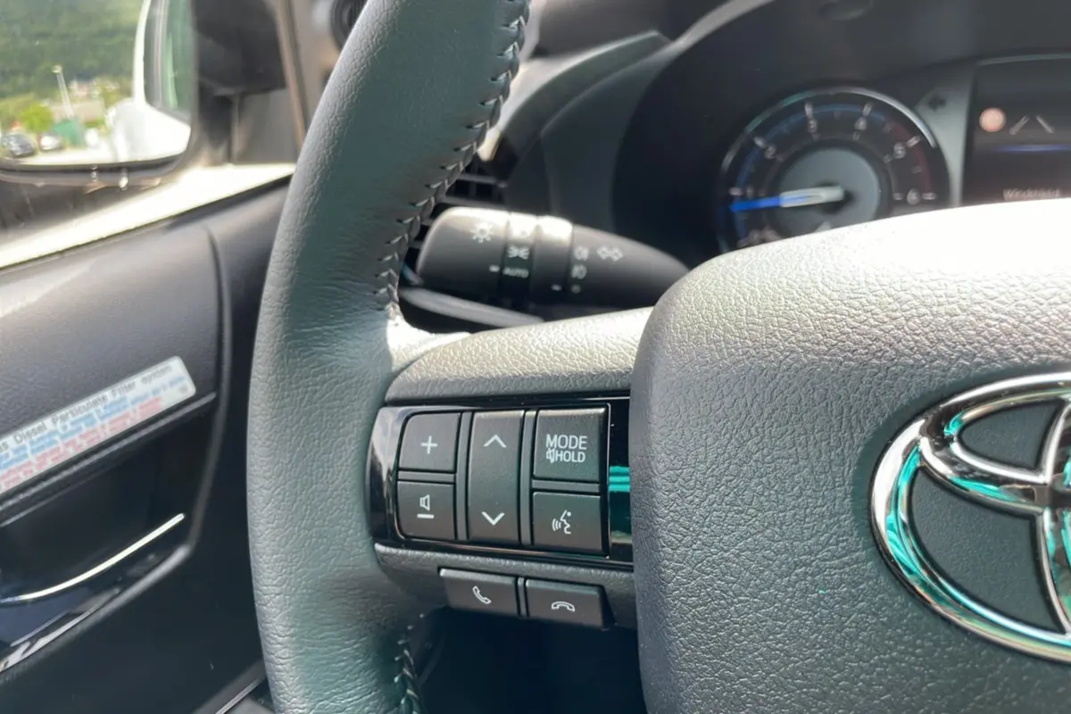 Gros plan sur le volant côté gauche du Toyota Hilux 2025 blanc, avec commandes audio et logo Toyota visible.