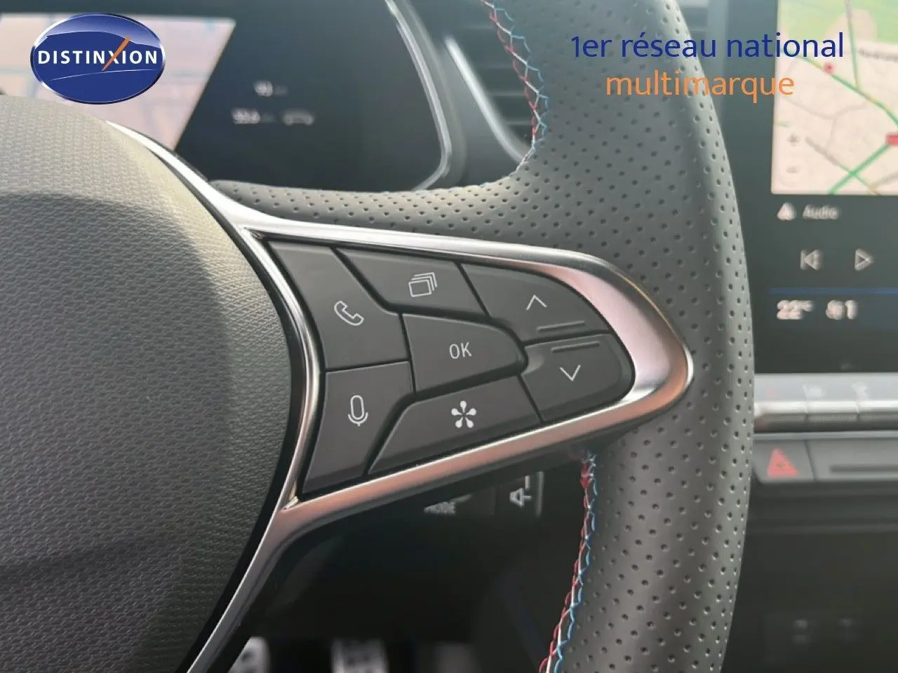 Vue rapprochée du volant en cuir perforé avec commandes multifonctions, tableau de bord et écran tactile en arrière-plan.