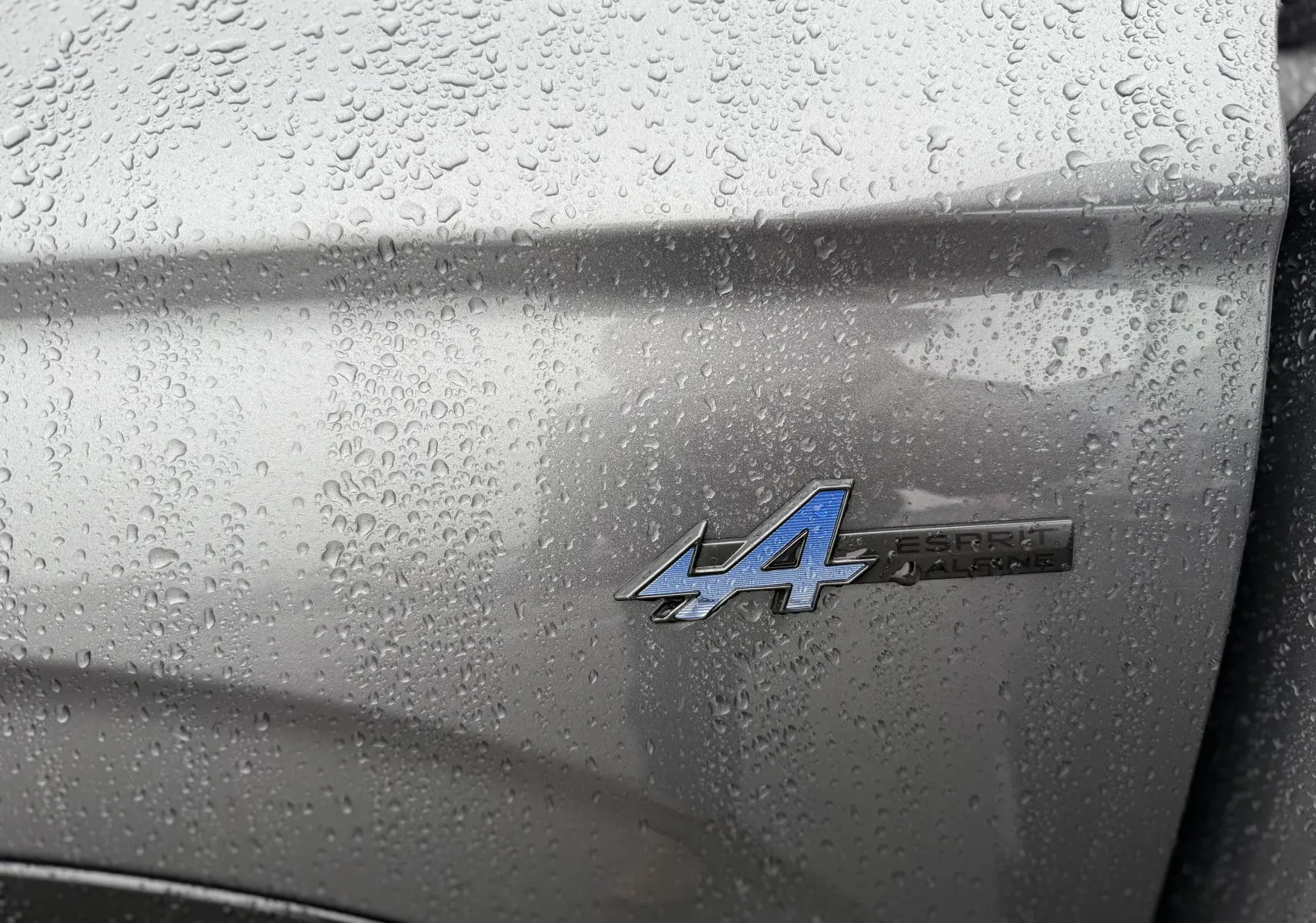 Gros plan sur l'écusson Esprit Alpine bleu sur la carrosserie gris schiste mouillée du Renault Austral 2025.